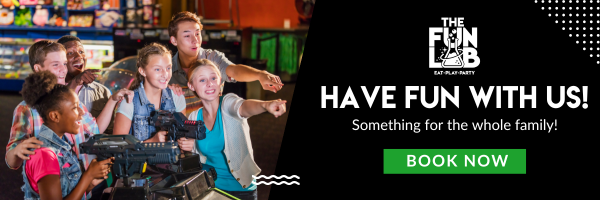 Family at arcade aiming game guns next to ad: THE FUN LAB, HAVE FUN WITH US!, Something for the whole family!, BOOK NOW
